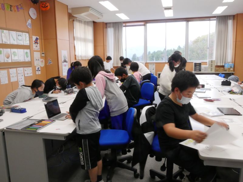 木島平小学校の教室で小学生の子ども達がパソコンや紙を使ってイラスト制作をしている様子の写真